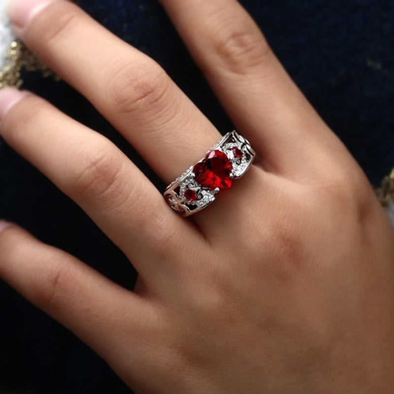 Heart-shaped engagement ring on finger with red stones and clear accents in silver-tone